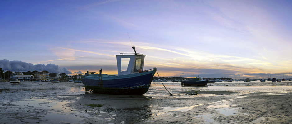 A small blue boat resting on wet sand at low tide with a calm harbor and colorful sunset sky in the background